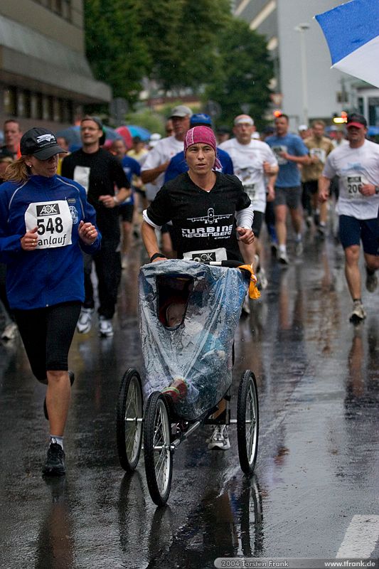 Läuferin des Teams "Turbine Heidenberg 03" mit Kinderjoggingwagen\n1. Siegerländer AOK – Firmenlauf am 9. Juli 2004 in der Siegener Innenstadt über 5,5 km mit über 2200 gemeldeten Teilnehmern. Die Witterungsbedingungen mit teilweise strömenden Regen und zurückhaltenden Temperaturen waren leider nicht dem sommerlichen Datum angemessen, was der Lauffreude jedoch keinen Abbruch tat.